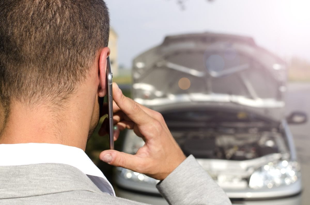 Automobiliste au téléphone devant sa voiture capot ouvert, appelant une assistance depuis le bord de la route